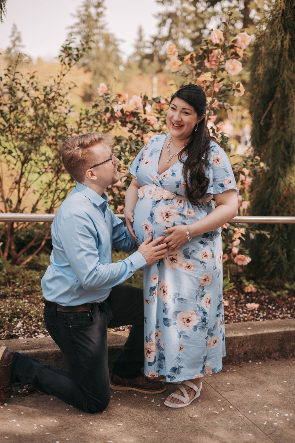Expectant father kneeling to touch pregnant woman's belly in floral blue maternity dress at garden park.