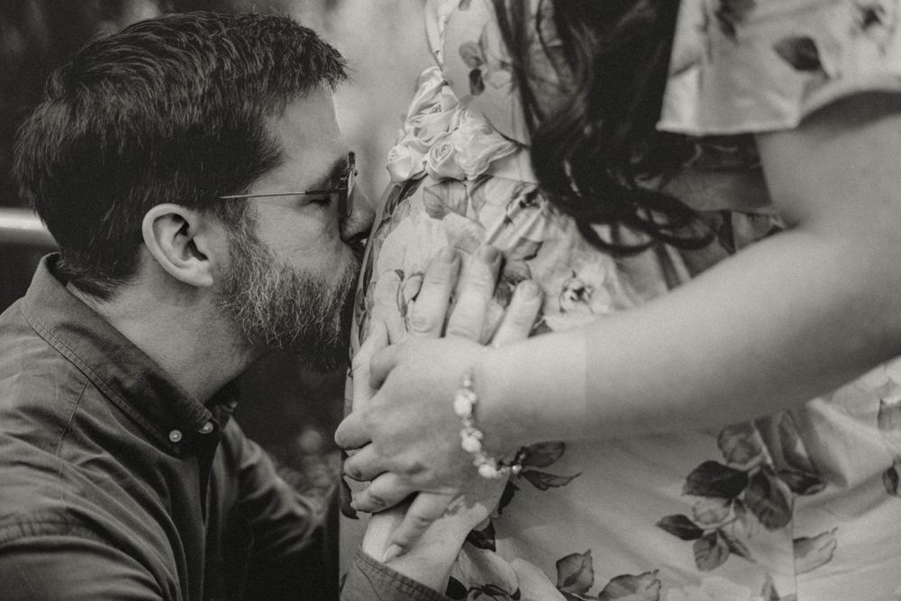 Black and white close-up of man kissing pregnant woman's belly while she holds his hands.
