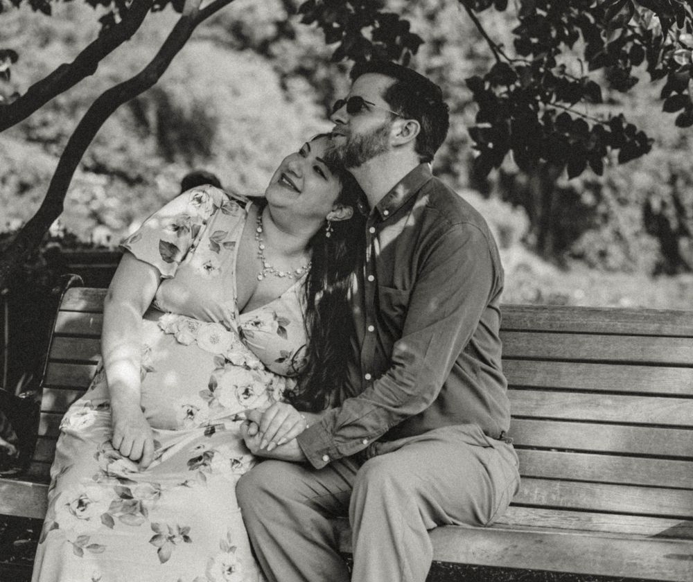 Black and white photo of couple sharing a kiss on park bench during maternity photoshoot.