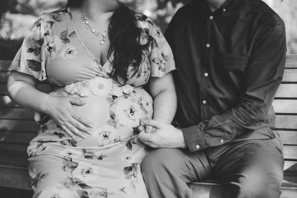 Black and white maternity photo of couple sitting on bench with hands on pregnant belly.