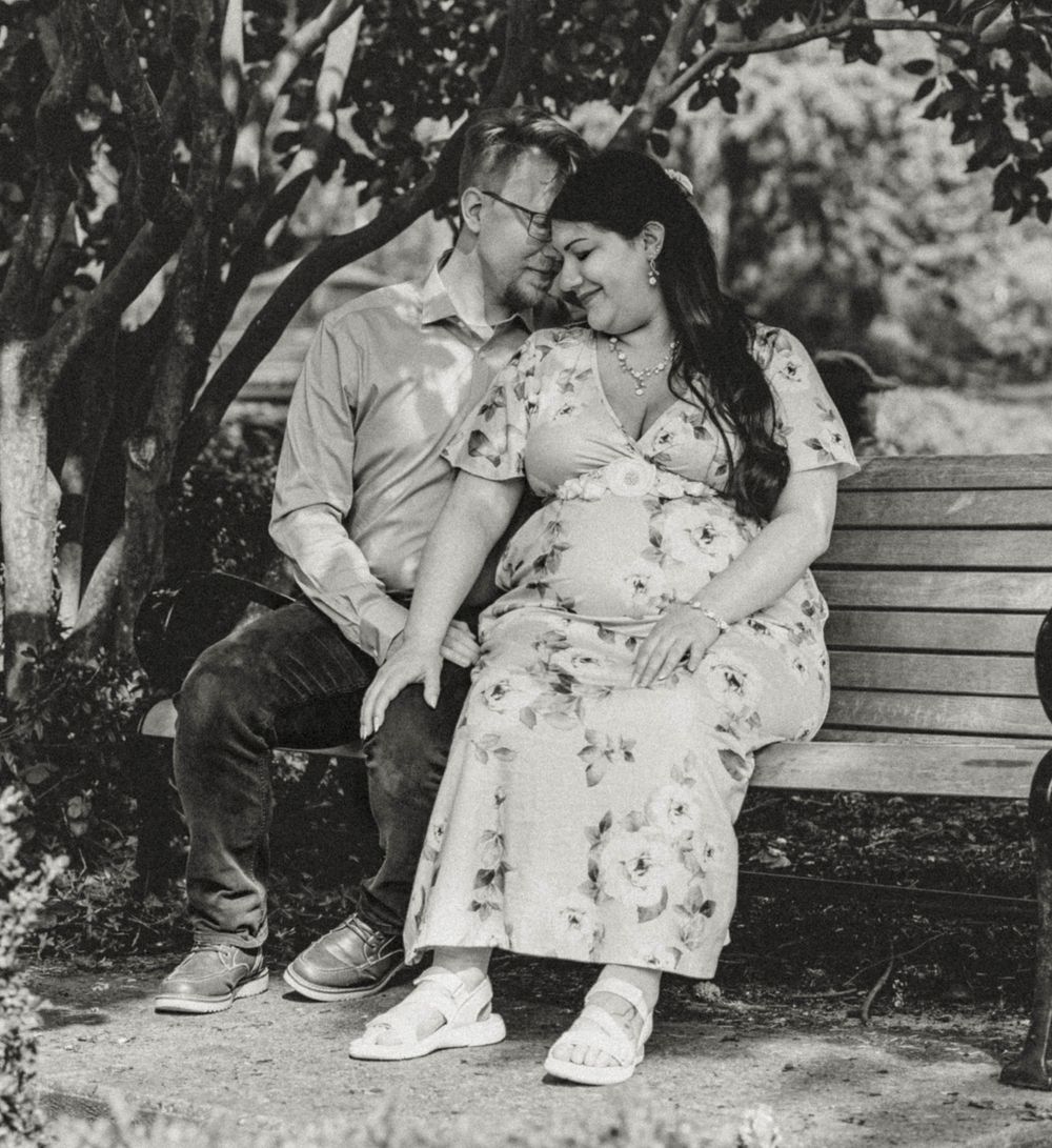 Black and white photo of couple sitting on park bench smiling at each other during maternity session.