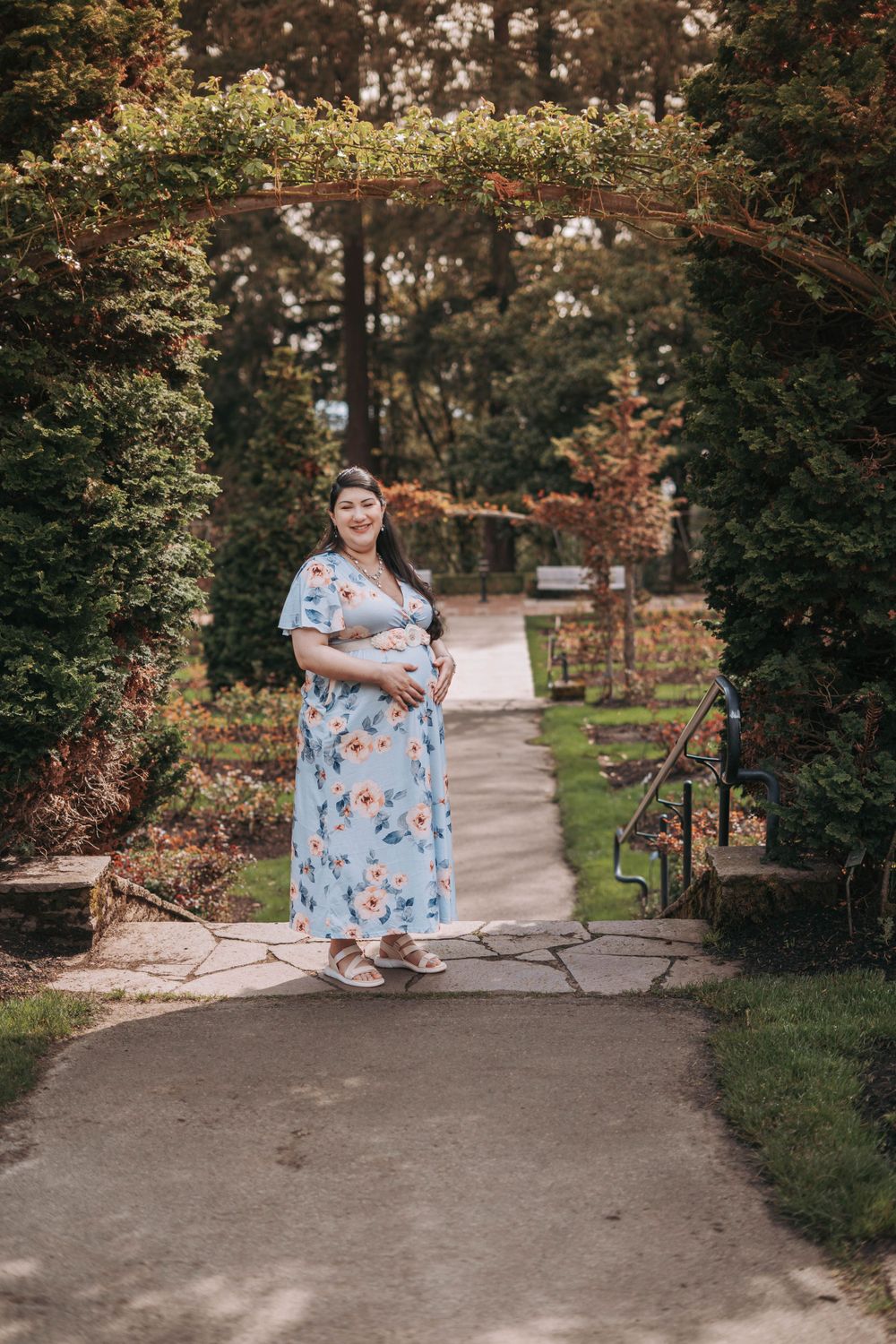 Smiling pregnant woman in light blue floral maxi dress standing under garden archway surrounded by greenery.