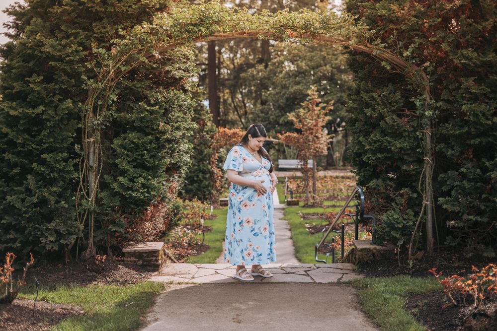 Pregnant woman in blue floral dress standing alone under lush garden arch on stone pathway.