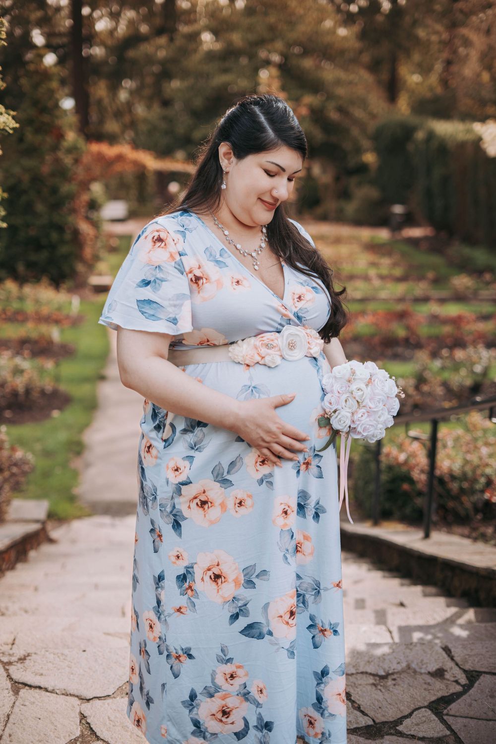 Pregnant woman in blue floral maxi dress holding baby bump and flowers in a garden.