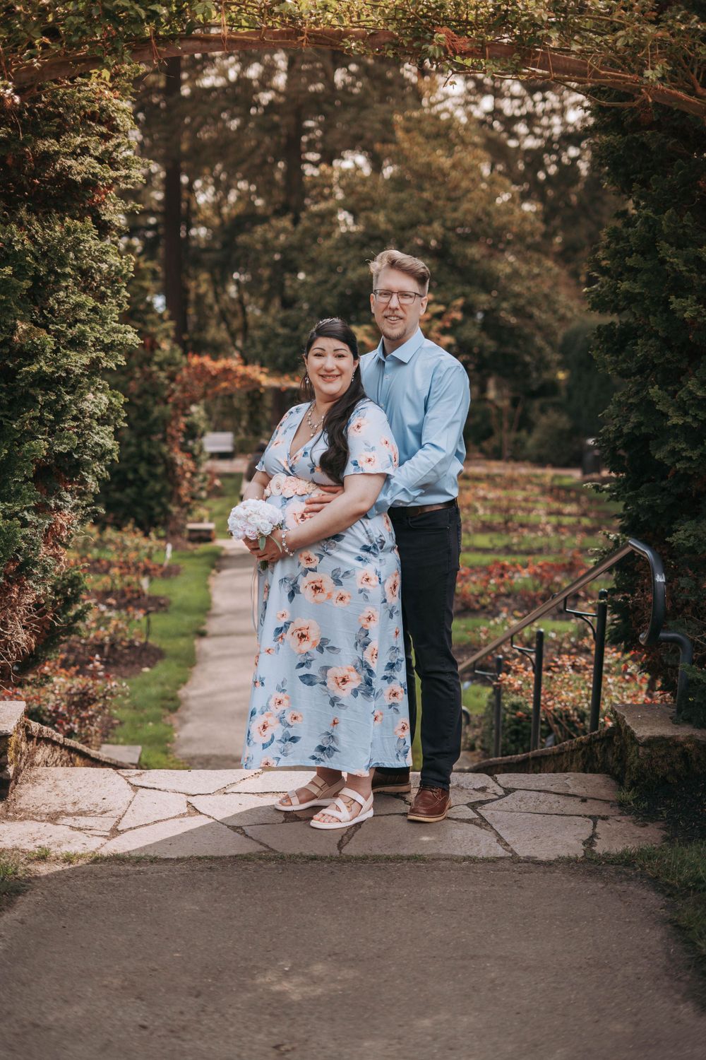 Happy couple posing under garden arch, pregnant woman in blue floral dress with partner in blue shirt.