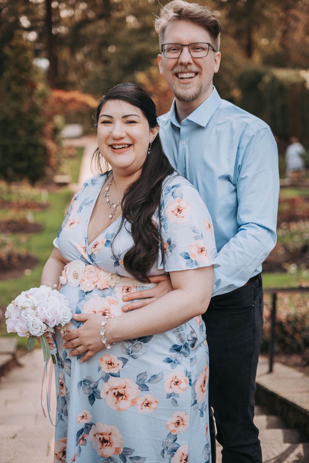 Smiling couple embracing outdoors, pregnant woman in blue floral dress holding white bouquet in garden.
