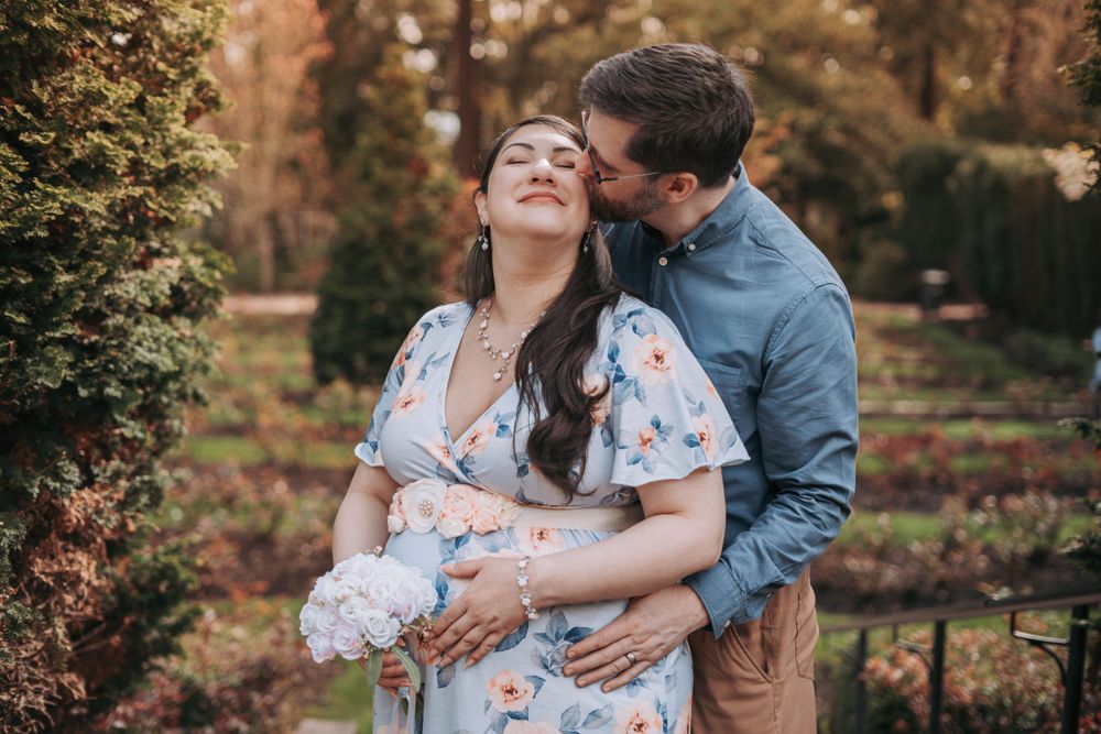 Man tenderly kissing pregnant woman's cheek while embracing her from behind in garden setting.