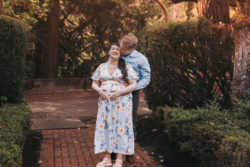 Couple embracing outdoors on brick path, woman in blue floral maternity dress in garden.