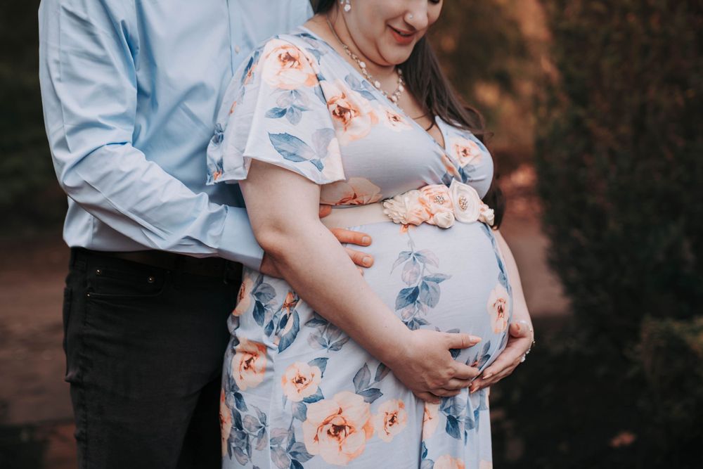 Close-up of pregnant woman in floral dress with partner holding baby bump outdoors.