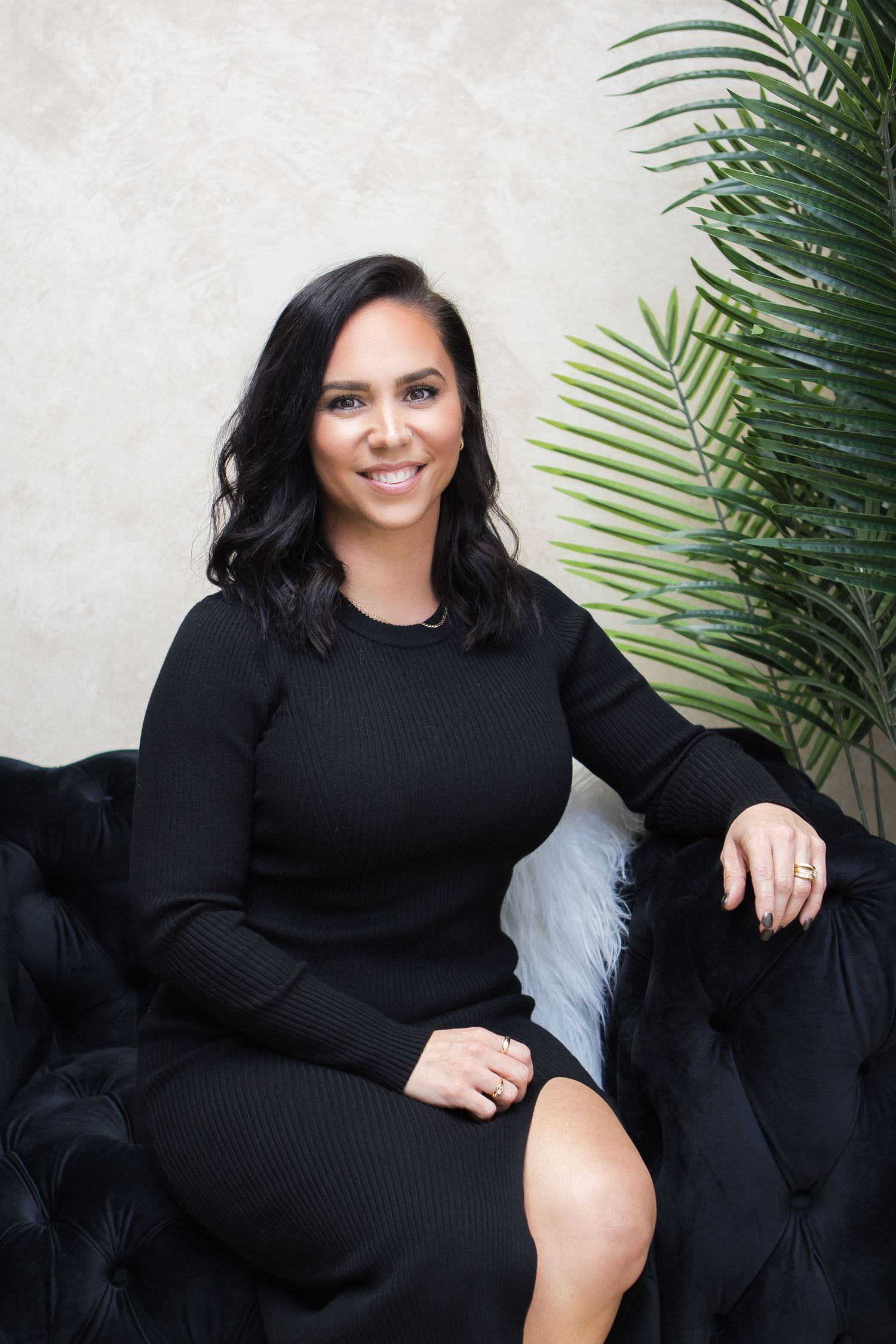 Woman in black sweater dress sits elegantly on dark couch with palm leaves in background.