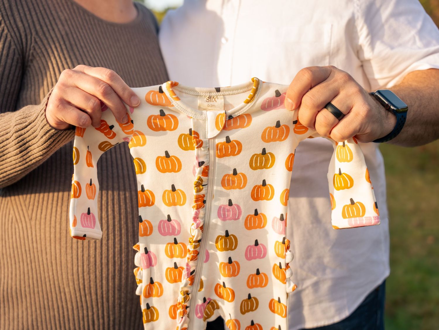 Close-up of hands holding festive pumpkin-printed baby clothes outdoors.