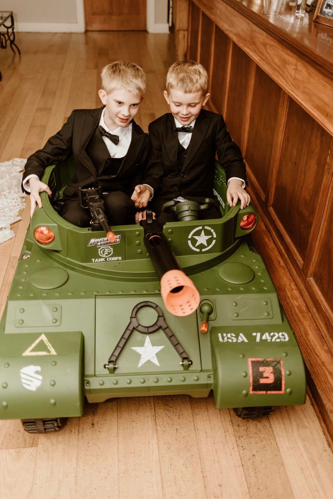 Two young boys in suits playing in a green toy military tank on a hardwood floor.