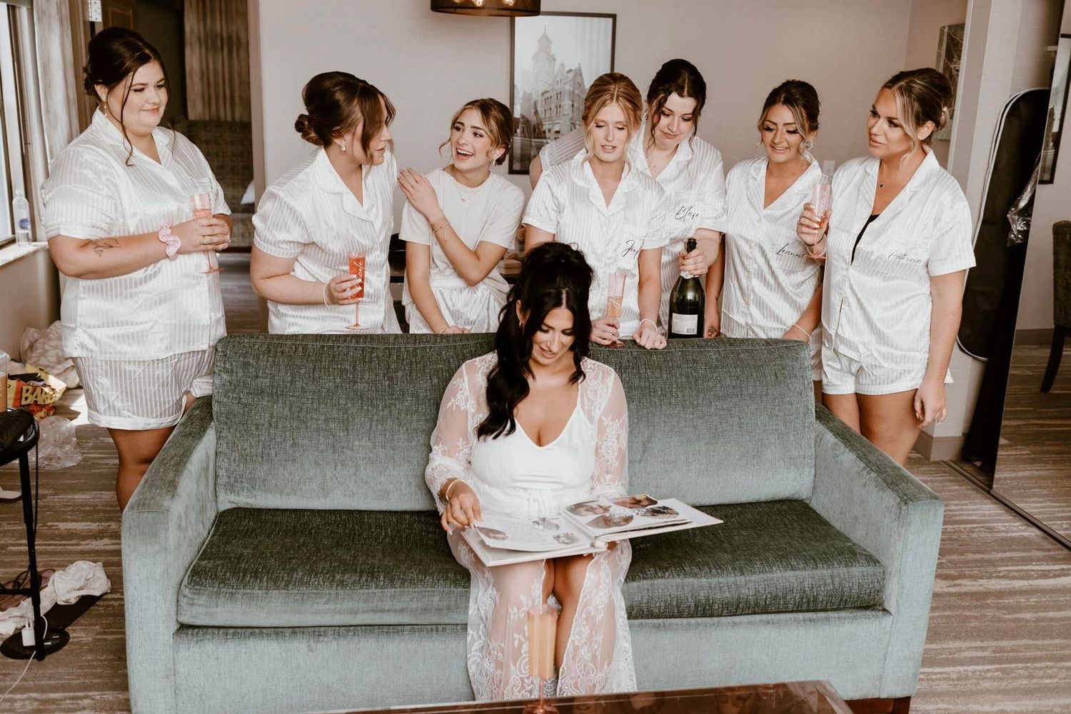Bridal party in white robes gathered around a gray couch during wedding day preparations.