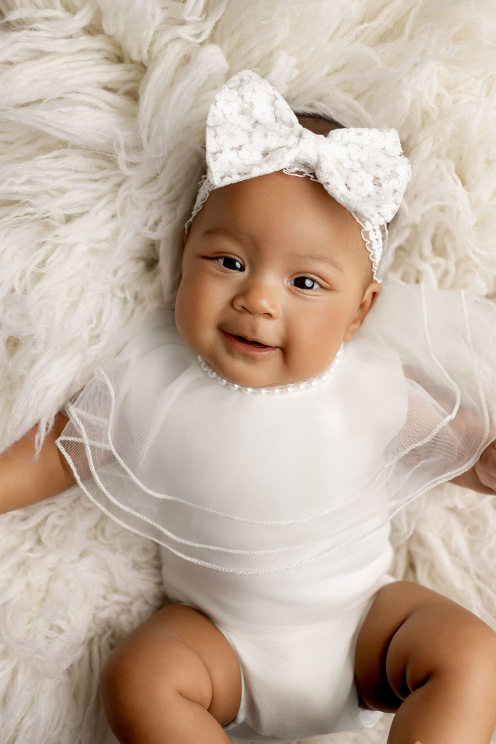 Happy baby wearing white lace bow headband and tulle outfit lies on soft fluffy cream fur blanket.