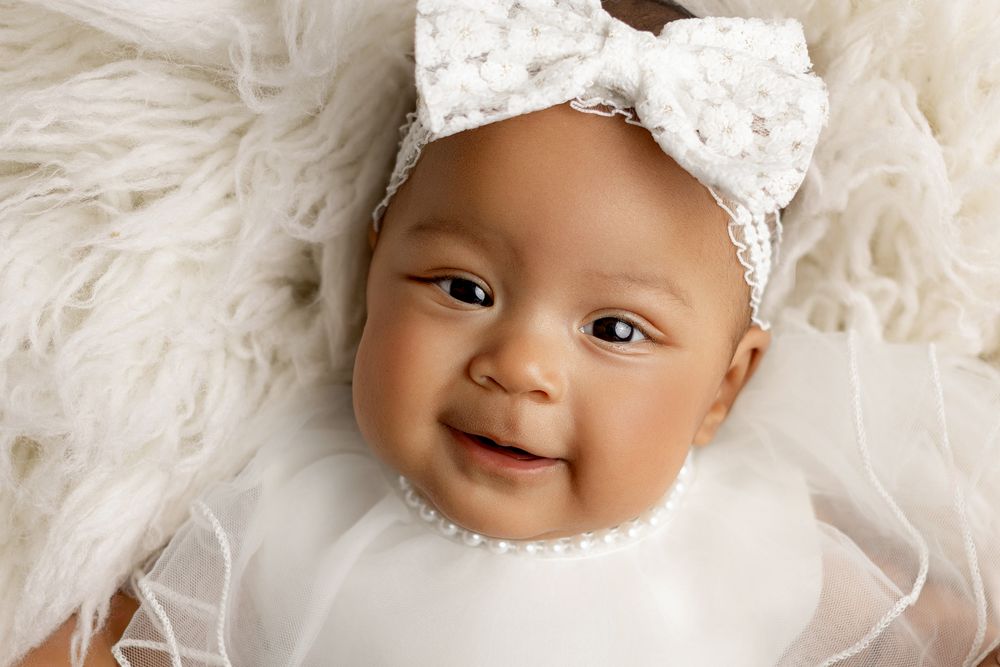 A smiling baby wearing a white lace bow headband lies on a soft white furry blanket.