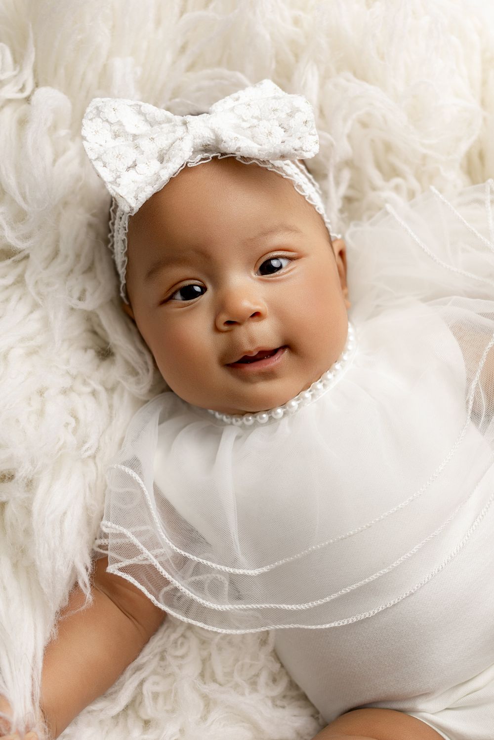 A smiling baby wearing a white lace headband bow lies on soft white fur with a white outfit.