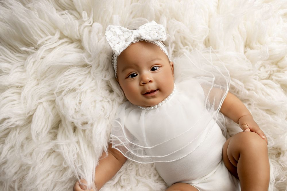 A baby wearing a white bow headband and bodysuit lies on a fluffy cream-colored fur blanket.