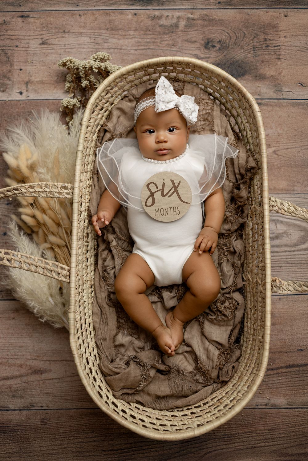 A baby wearing a white bow and onesie lies in a woven bassinet with dried flowers and textured fabric.