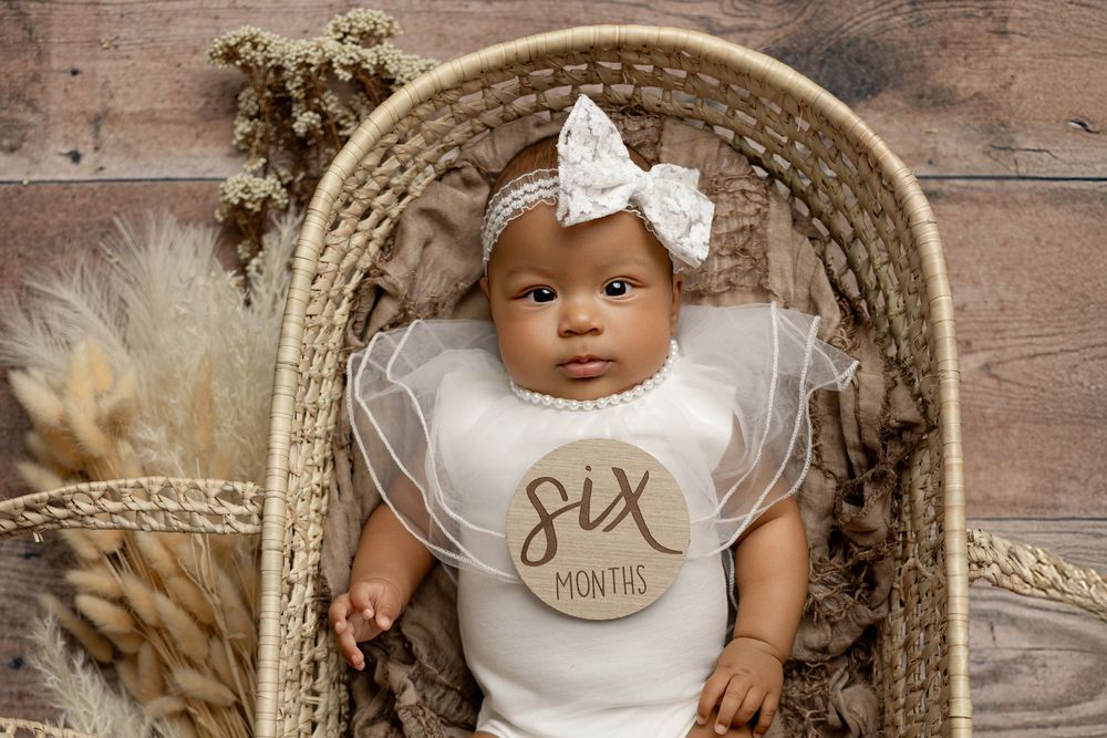 Baby in white outfit with bow headband lying in woven wicker basket with dried floral decorations.