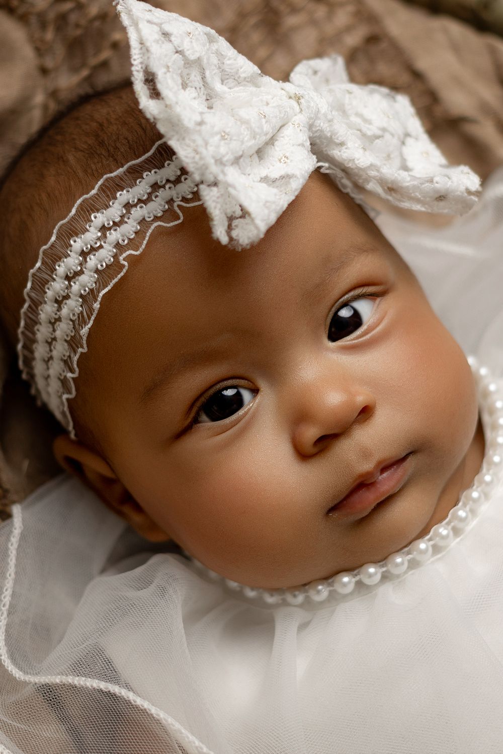 Small baby wearing white lace headband with bow and white dress gazes at camera with bright eyes.
