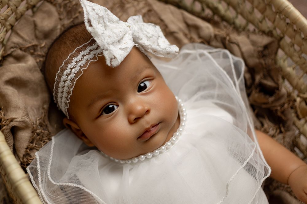 Infant wearing white dress and headband with bow lies on brown blanket.