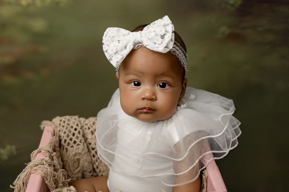 A small baby wearing a white tulle dress and lace bow headband poses in a wicker chair.