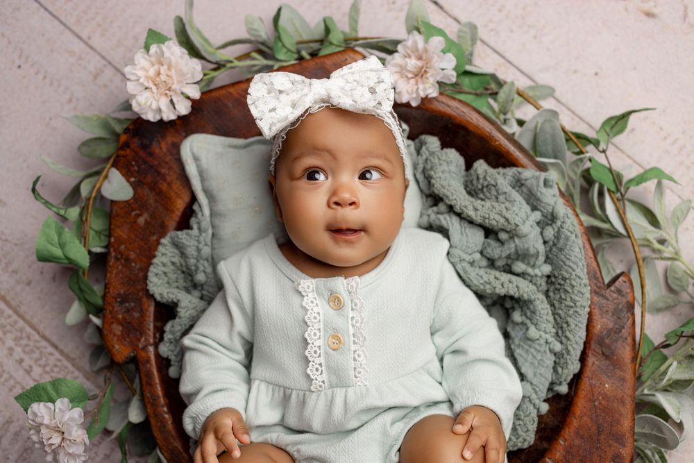 Baby wearing mint green outfit and white bow lying on textured blanket with floral decor.