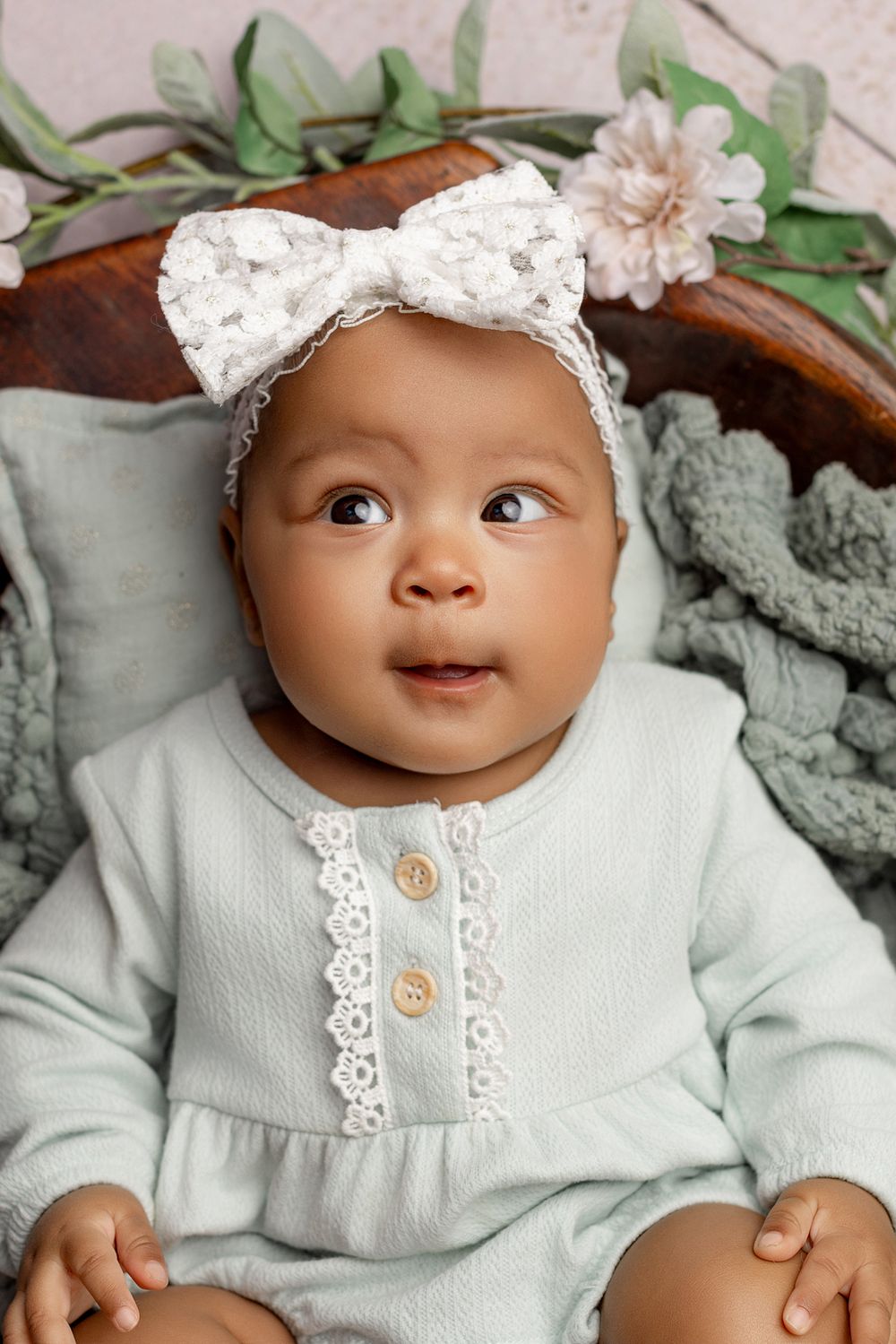 Adorable baby in mint green lace-trimmed outfit with white bow headband lying on cushions with floral decorations.