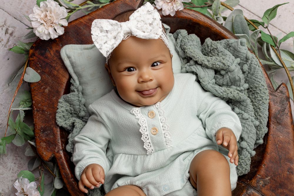 Baby in mint colored outfit smiling while resting on grey blanket with decorative greenery.
