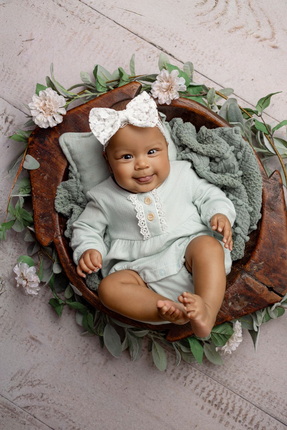 Baby in mint green outfit lying in wooden bowl decorated with flowers and greenery.
