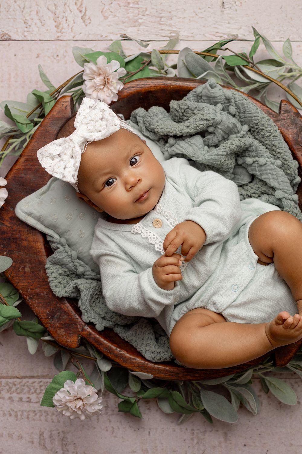 A baby in white clothing rests in a wooden bowl surrounded by greenery and gray blankets.