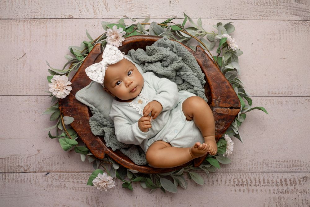 Baby wearing mint outfit rests in wooden bowl with greenery and floral decorations on gray background.