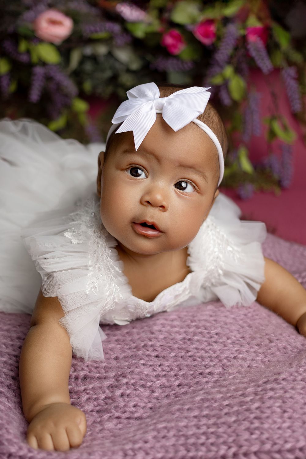 Baby in white dress and bow headband on purple knit blanket with floral background.