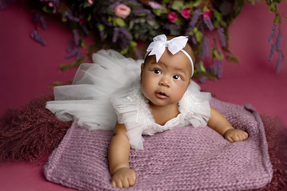 Baby in white dress and bow lying on purple pillow against pink background with floral decor.