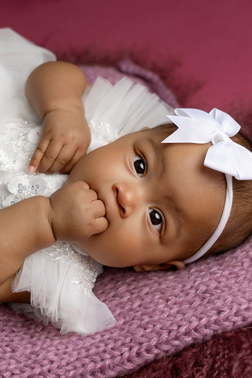 A baby in white lace dress and bow headband laying on pink knitted fabric.
