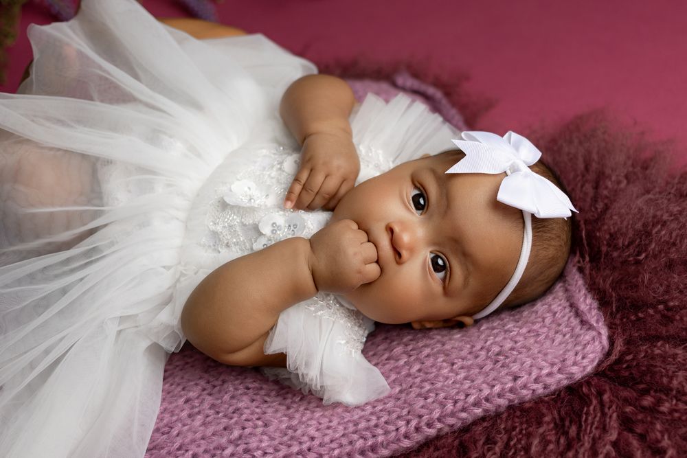 Baby in elegant white tulle dress and bow headband reclining on pink textile.
