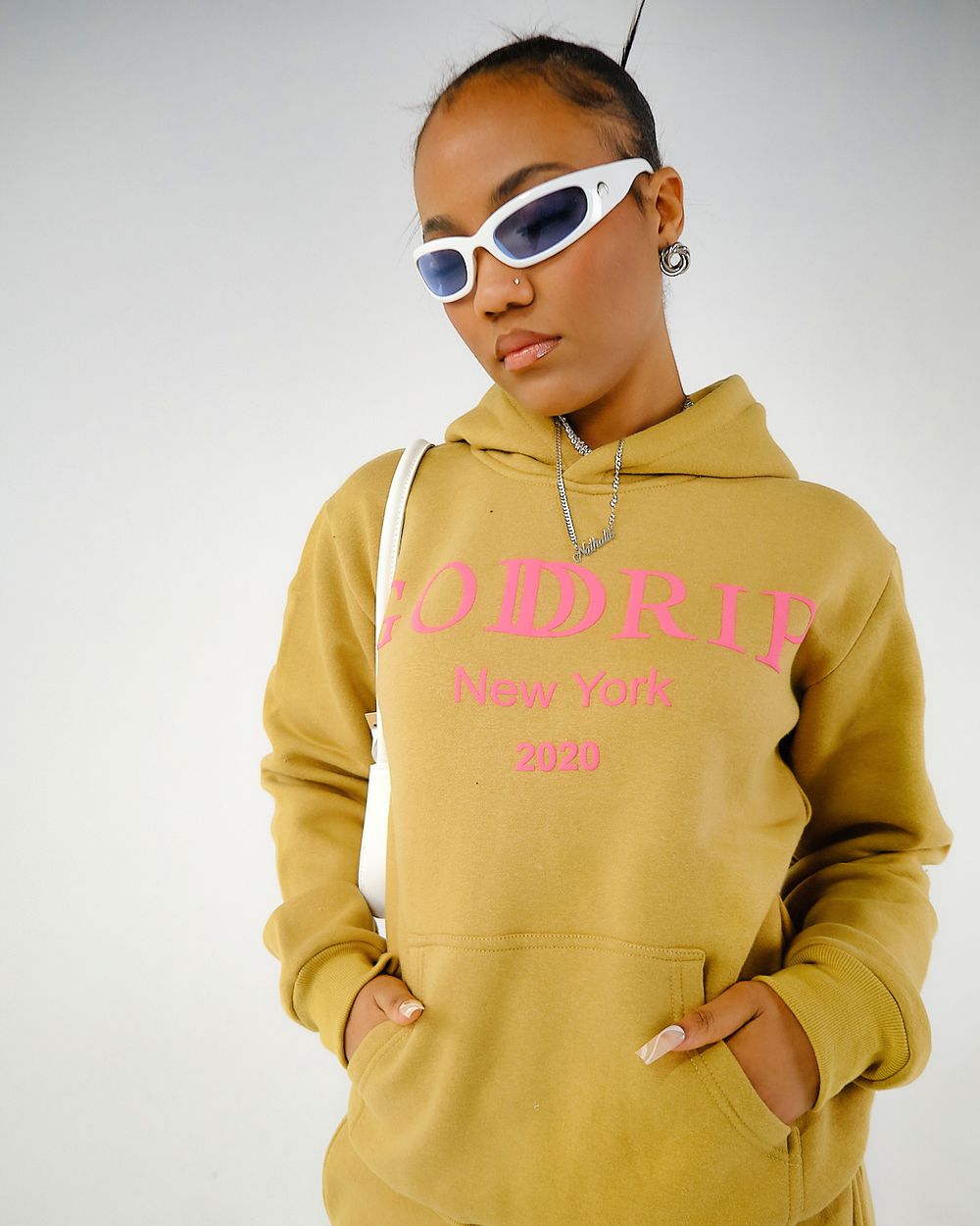 Model in mustard yellow hoodie with pink text and white sunglasses poses against light background.