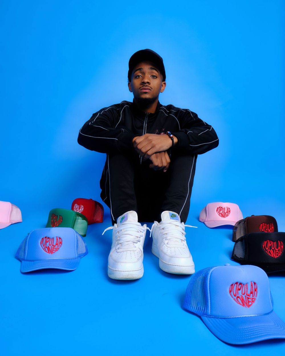 Person in black tracksuit sitting with white sneakers surrounded by colorful baseball caps on blue background.