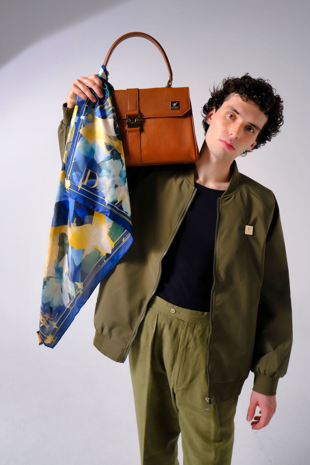 A model wearing an olive green workwear outfit holds a colorful scarf and brown leather bag.