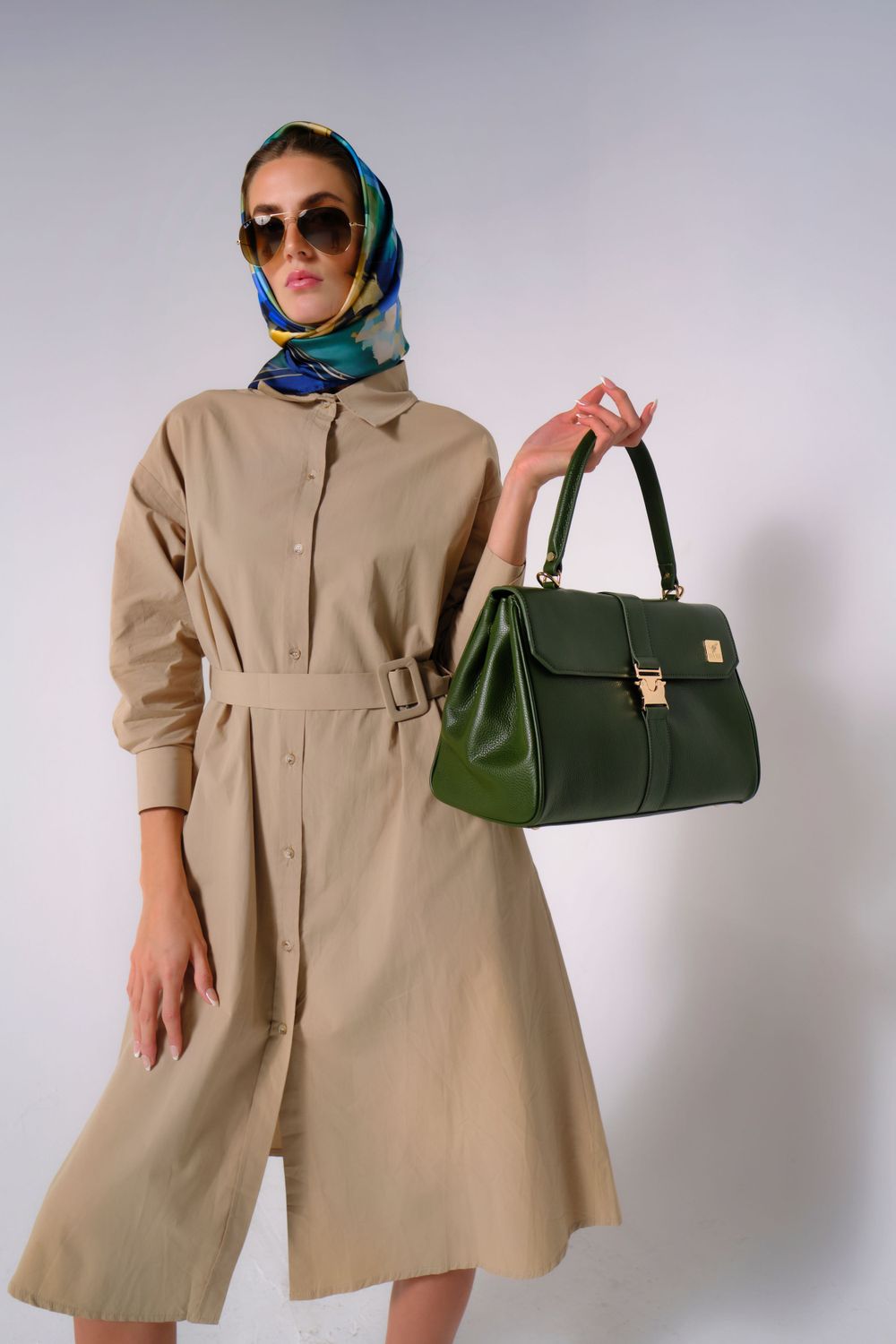 Model in beige trench dress and blue headscarf poses with green structured handbag.