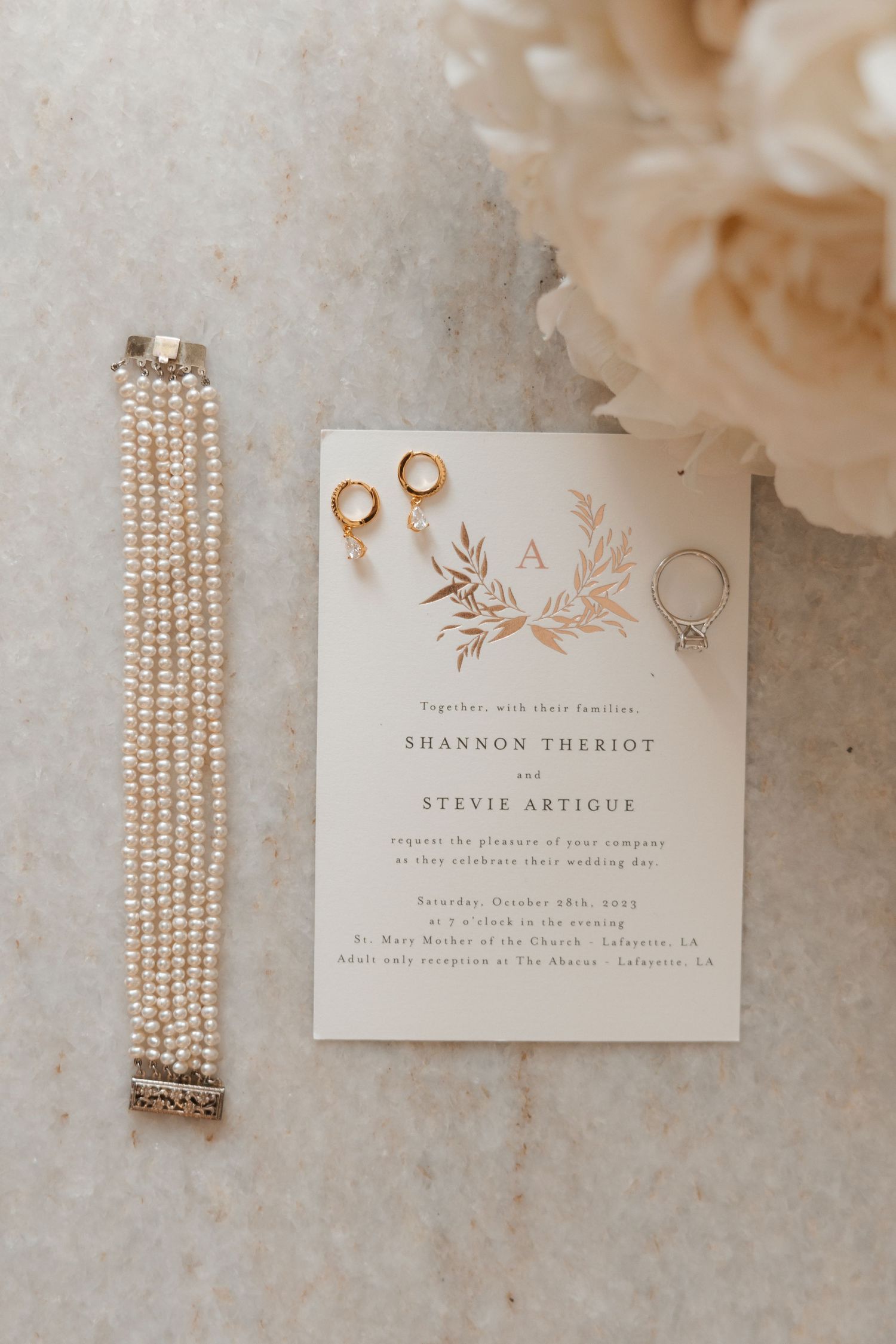 Elegant wedding invitation with gold foil details displayed alongside pearl bracelet and rings on marble surface.