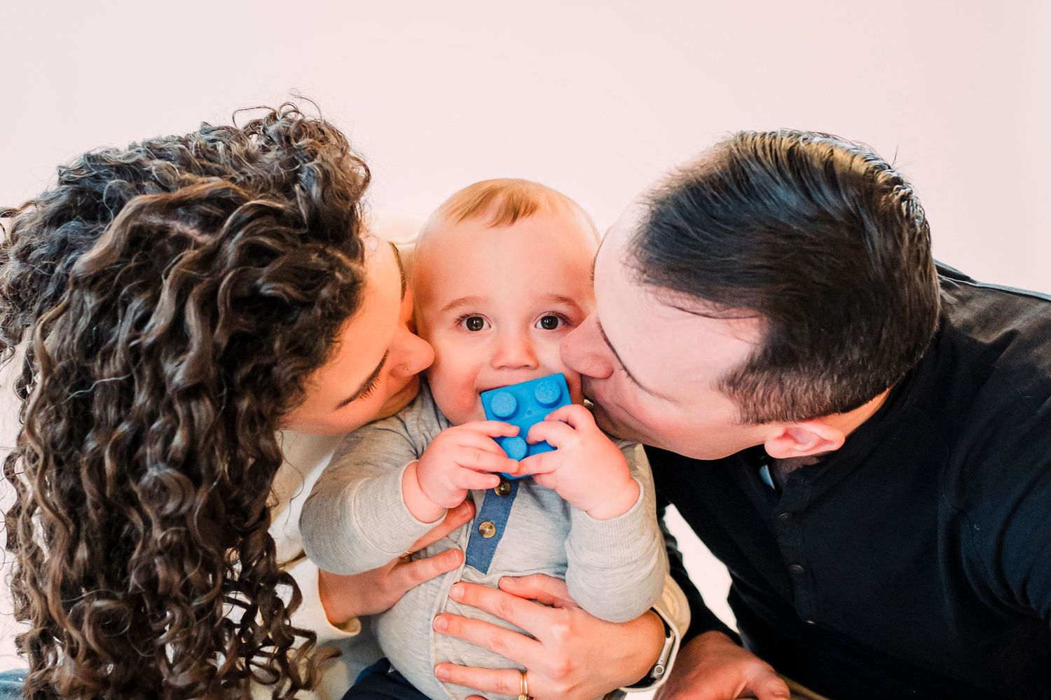 A baby with a blue pacifier receives kisses from loving parents against a light pink background.