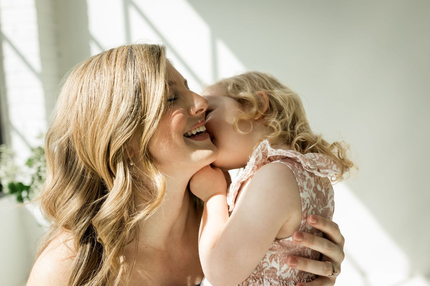 Mother laughing while young daughter kisses her during motherhood photography session in a studio in Calgary
