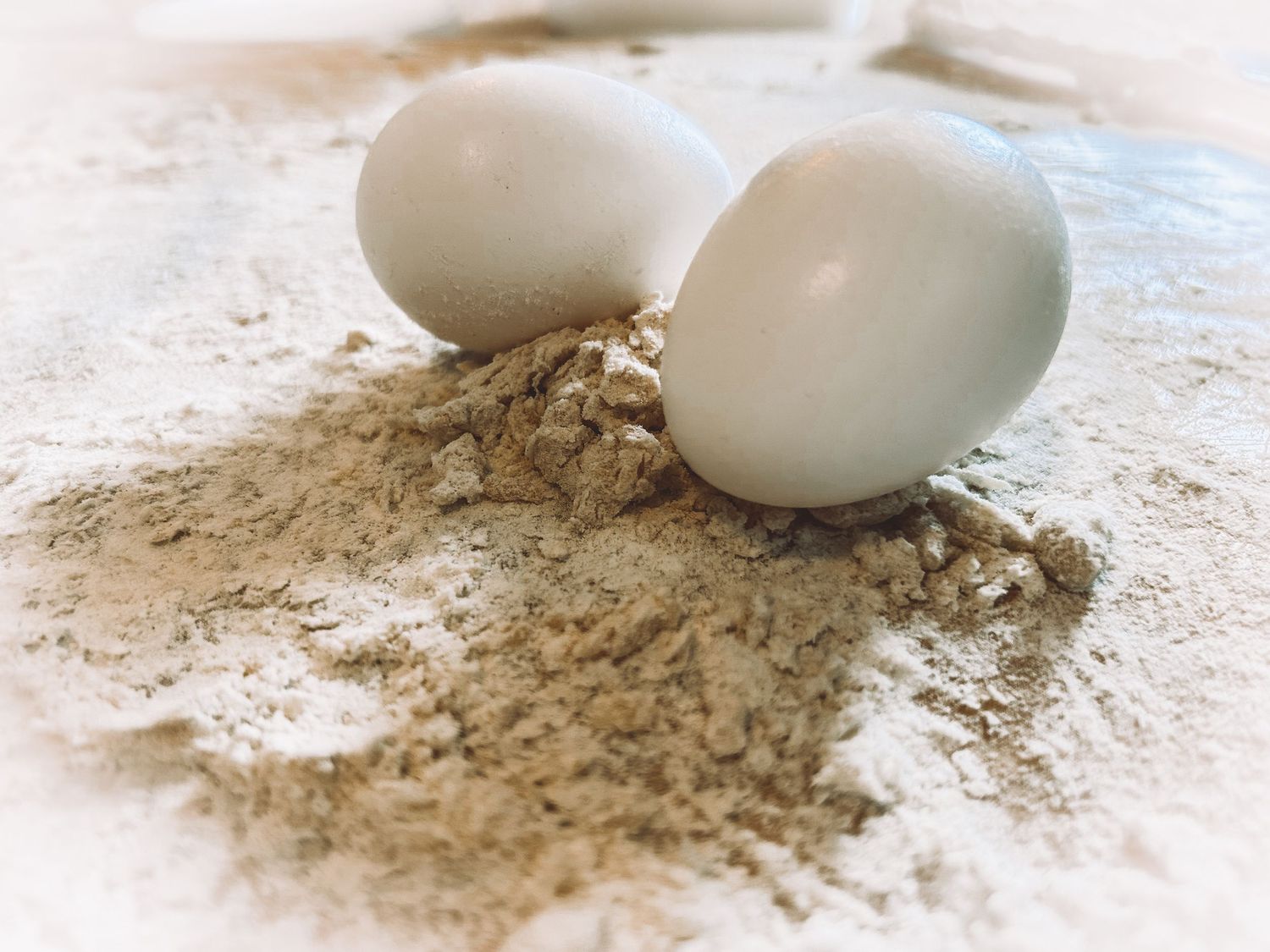 Two white eggs resting on a pile of flour with scattered grains on a light surface.