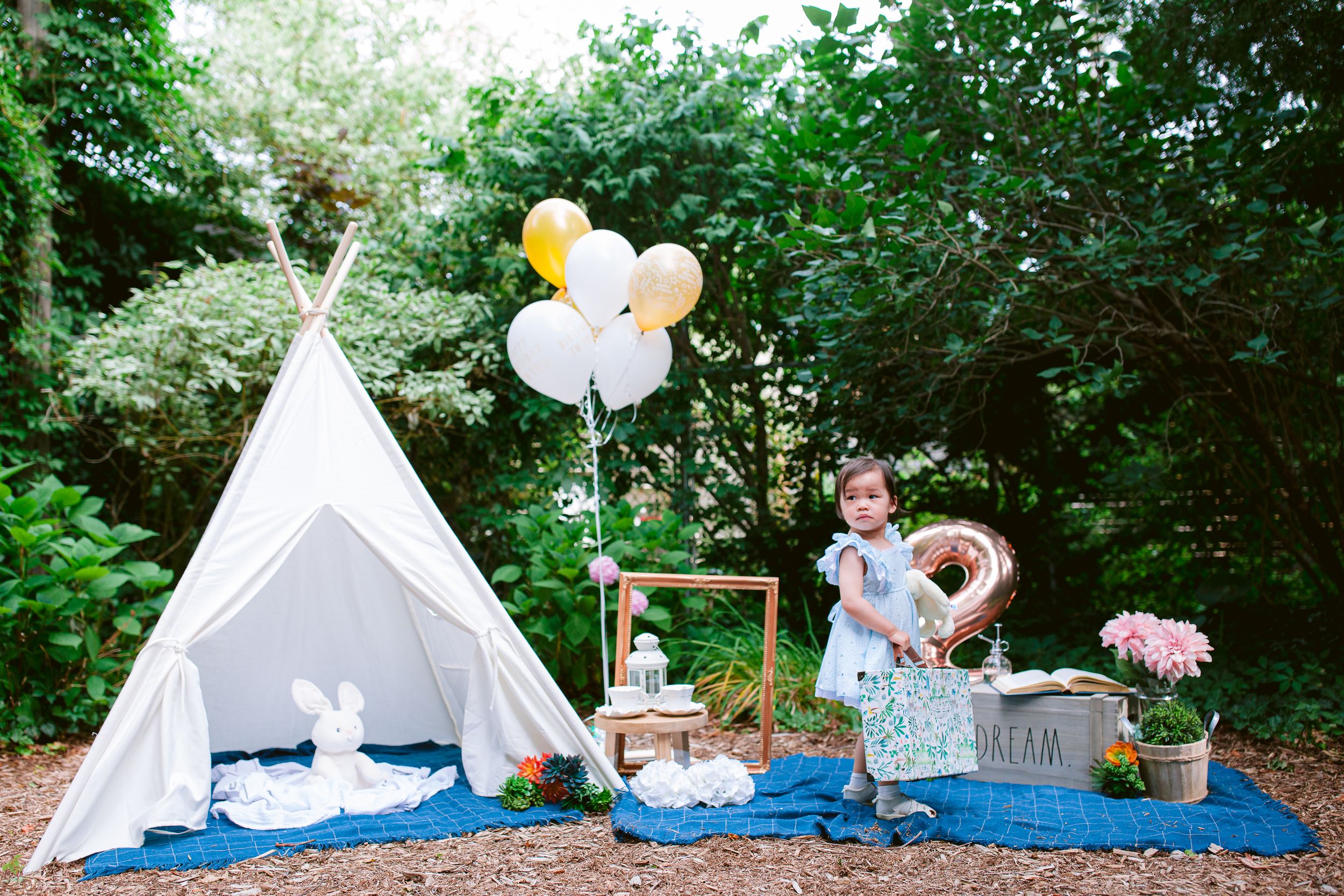 Whimsical outdoor party setup with white teepee tent, blue blanket, balloons and decorative party accessories in garden setting.