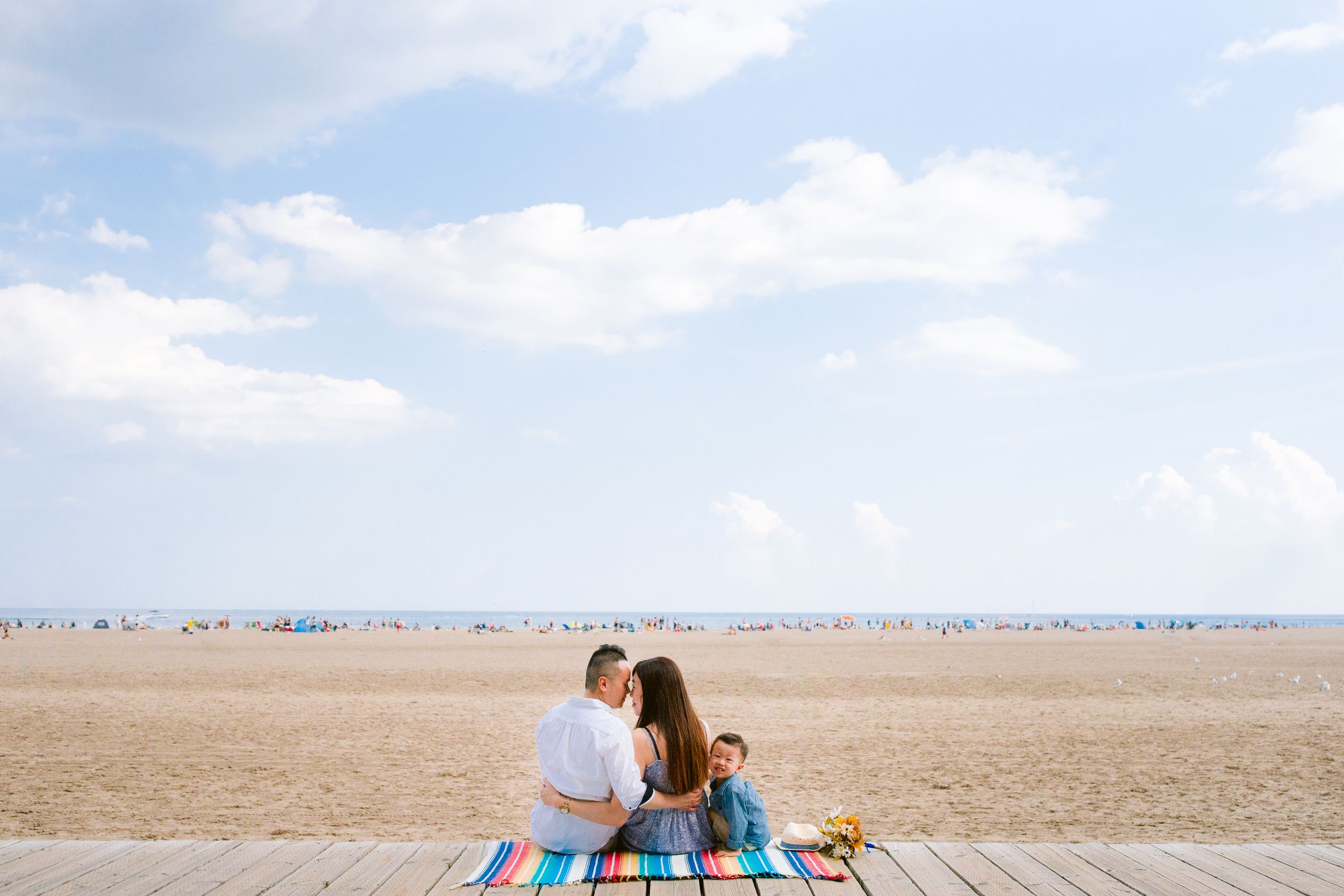 Two people share a tender moment on a blanket at an empty beach boardwalk overlooking the sand and horizon.