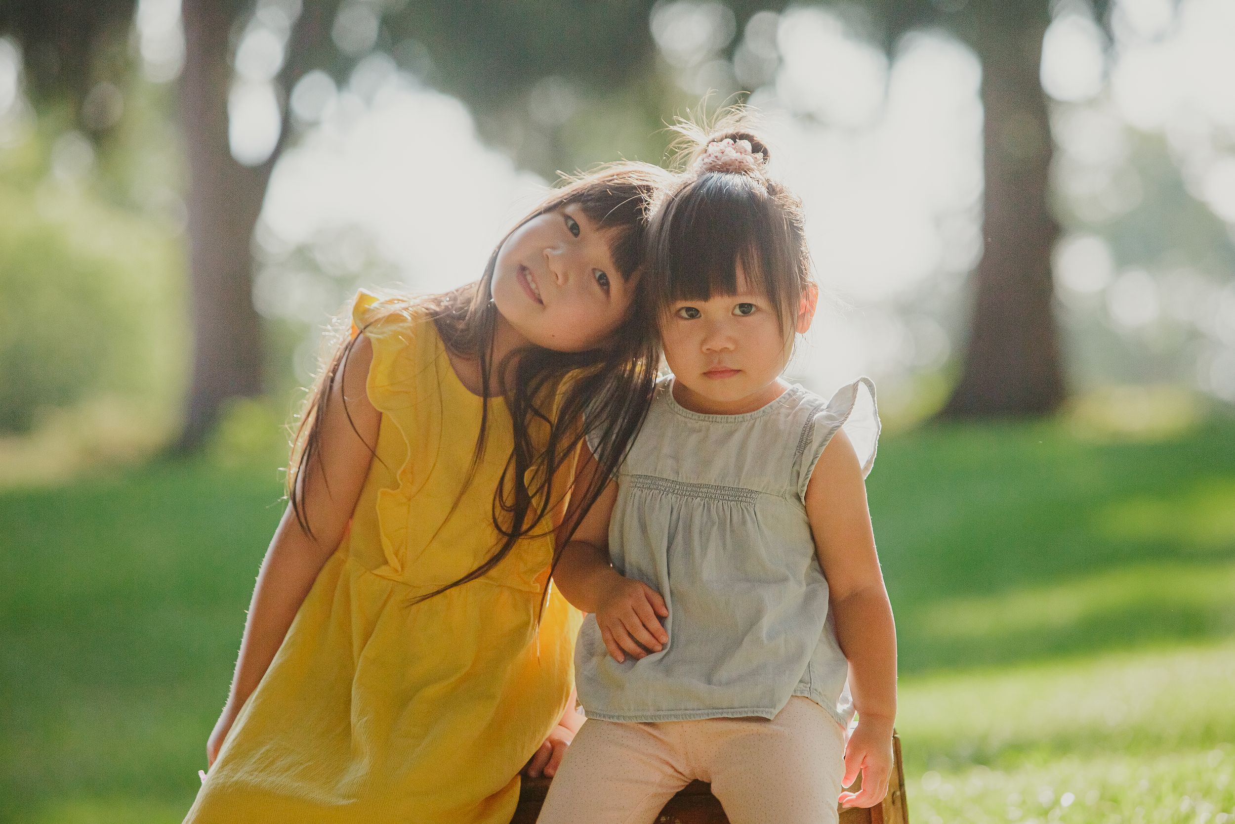 Two children in yellow and grey dresses lean together while sitting outdoors in a sunny park setting.