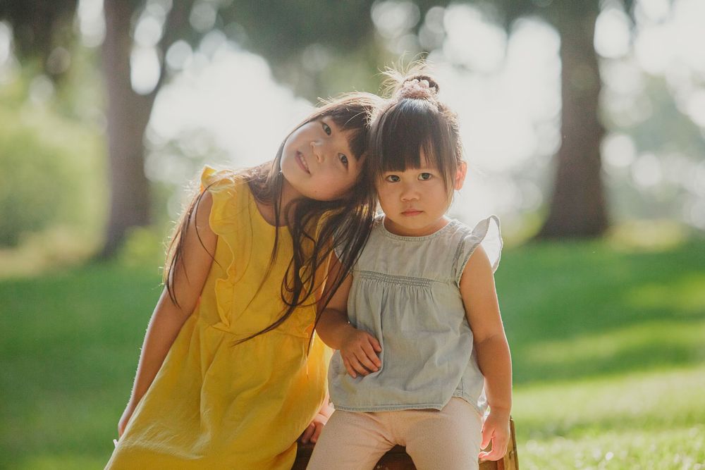 Two children in yellow and grey dresses lean together while sitting outdoors in a sunny park setting.