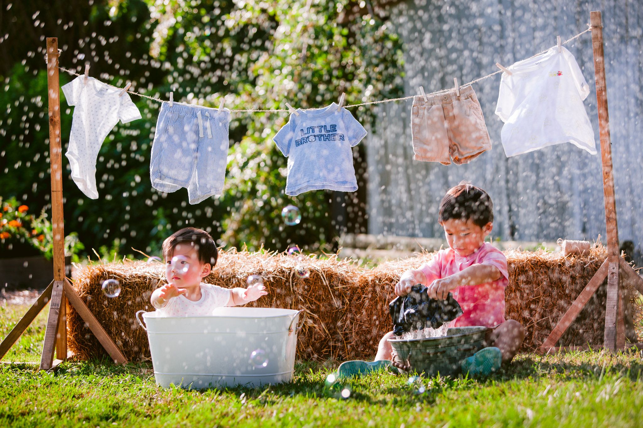 Two children play and splash in water basins on a summer day with laundry hanging on a clothesline in the background.