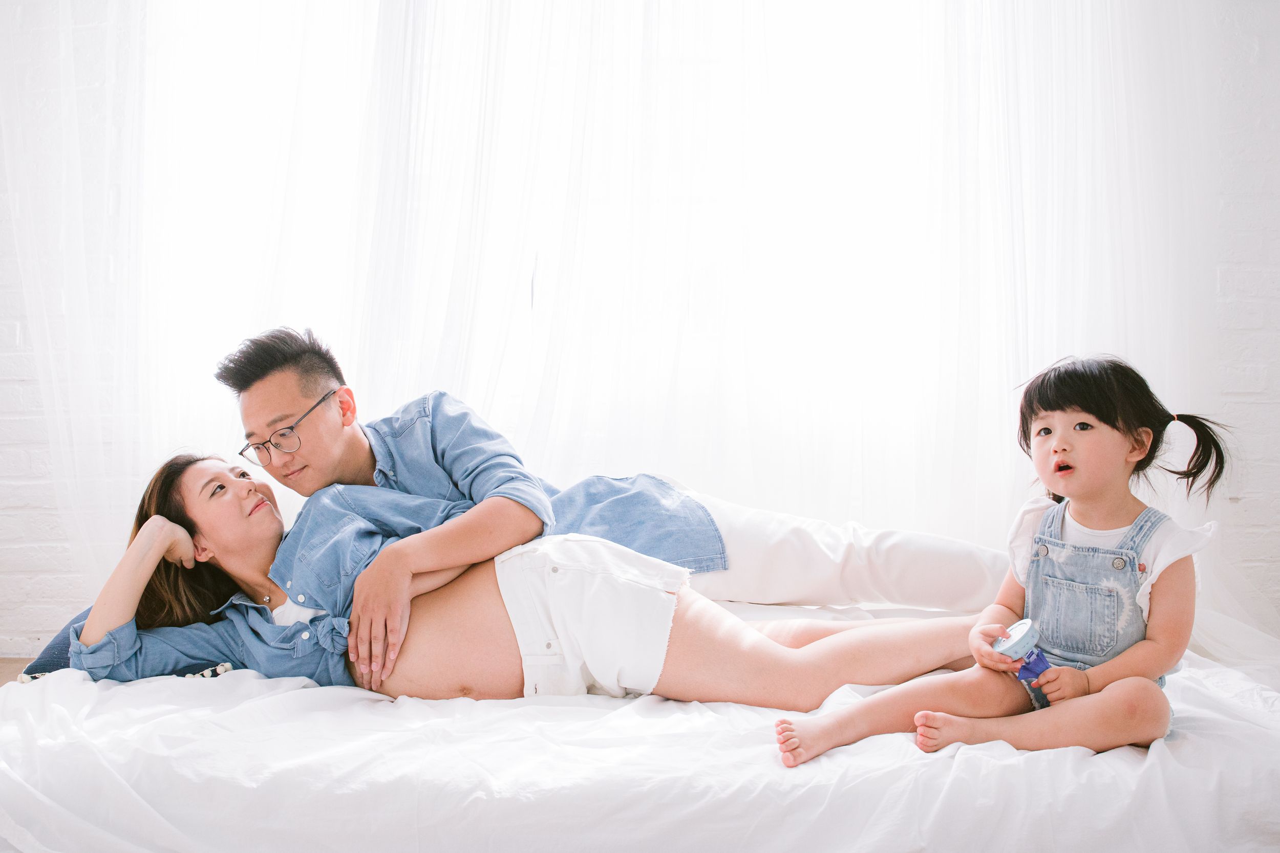 Family in casual attire relaxes together on white bedding, with small child sitting at their feet.